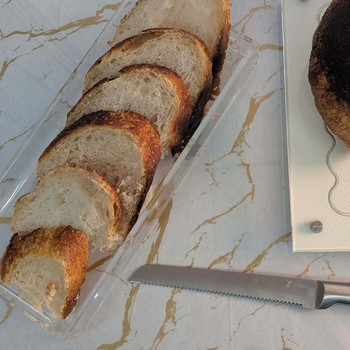 Sliced artisanal bread in a clear lucite bread tray on a marble-patterned surface, with a serrated bread knife and a white cutting board with a burnt piece of bread - Zstander