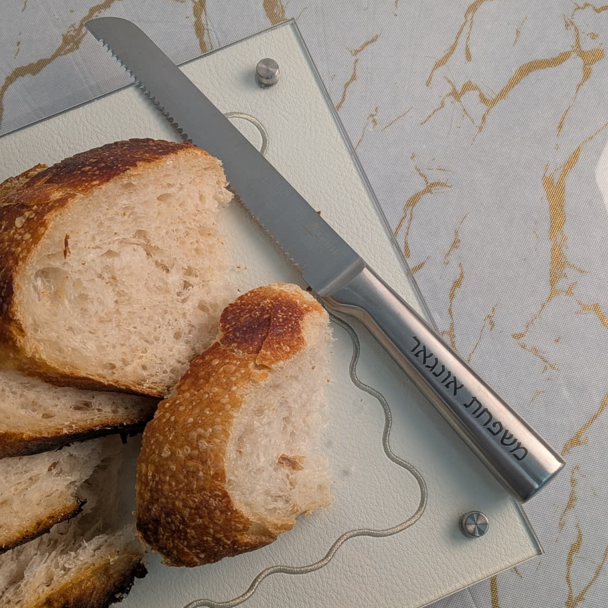 A serrated bread knife with a silver blade and a metallic handle engraved resting on a white wavy-edged cutting board with sliced bread on a marble-patterned surface - Zstander