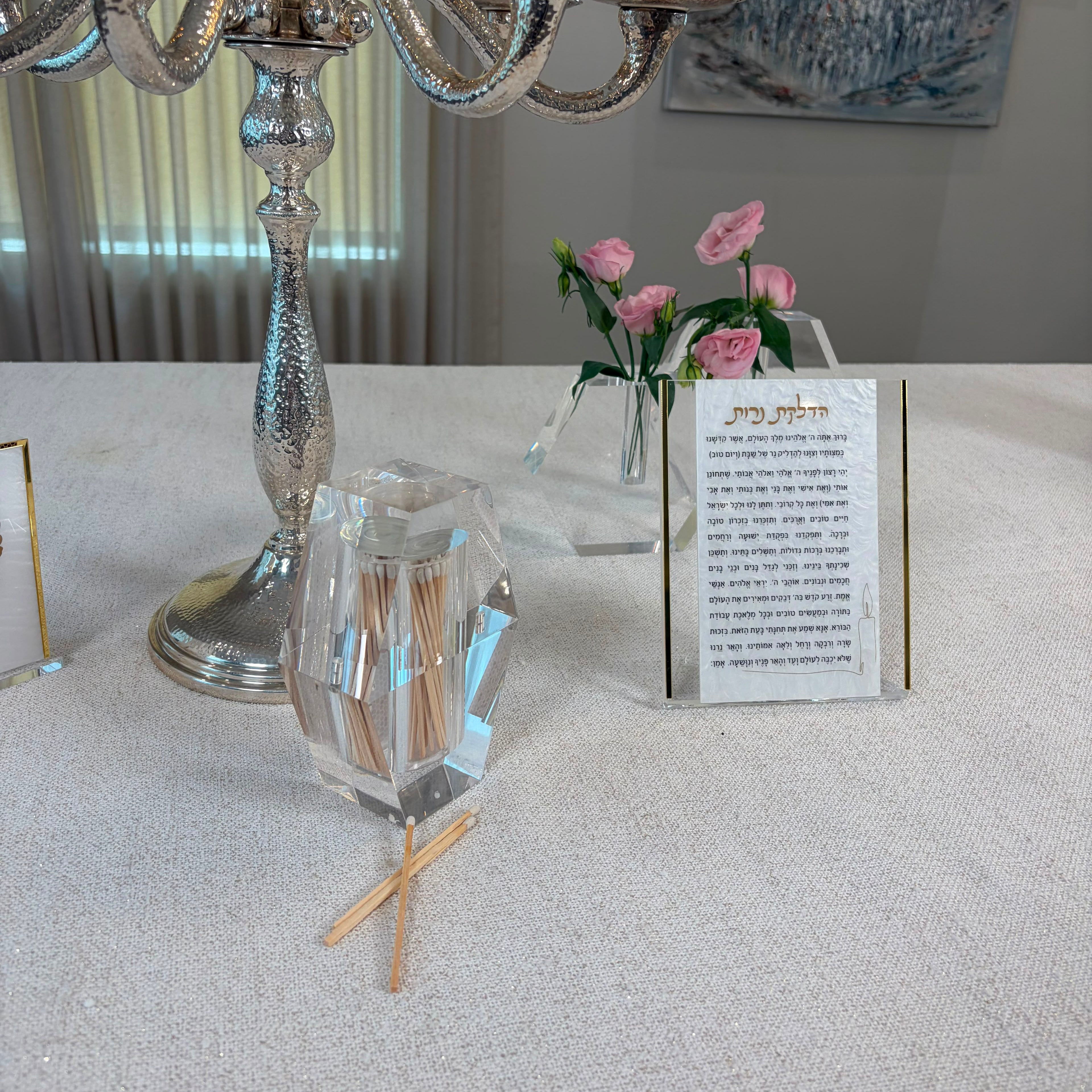A silver candelabra and a clear faceted glass match holder with wooden matches on a light beige tablecloth, alongside a small card with Hebrew text and pink flowers in a vase - Zstander