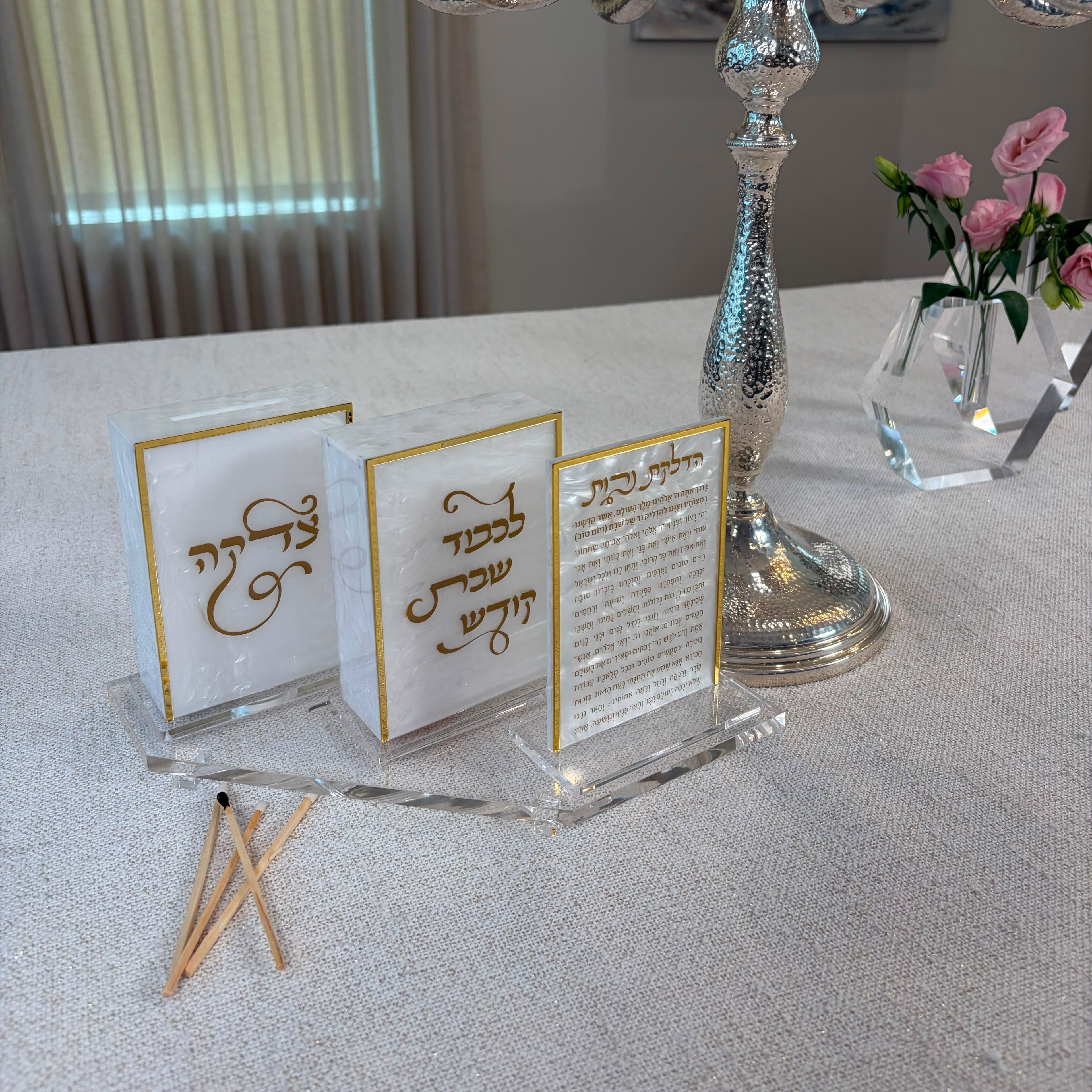 a clear tray with a hadlokas neiros card, tzedakah box and match box next to silver candlesticks on a table -Zstander