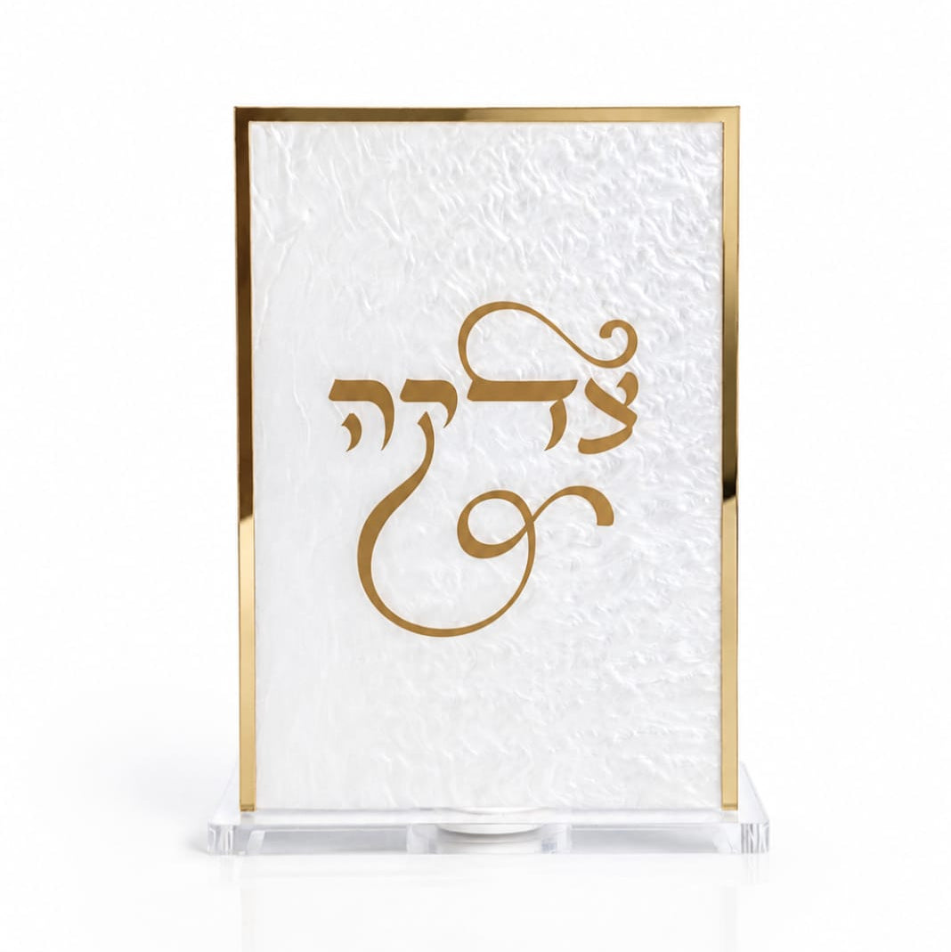 A rectangular pearl white Tzedakah box with a metallic gold border and a gold Hebrew script design in the center, displayed on a clear acrylic stand against a white background - Zstander