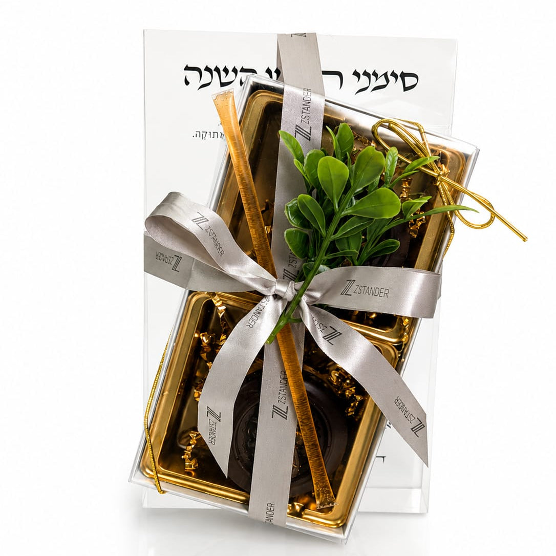 A gift box of chocolate wrapped in a silver ribbon with a green leafy sprig, set against a white background with a Rosh Hashana simanim Prisim block behind it - Zstander