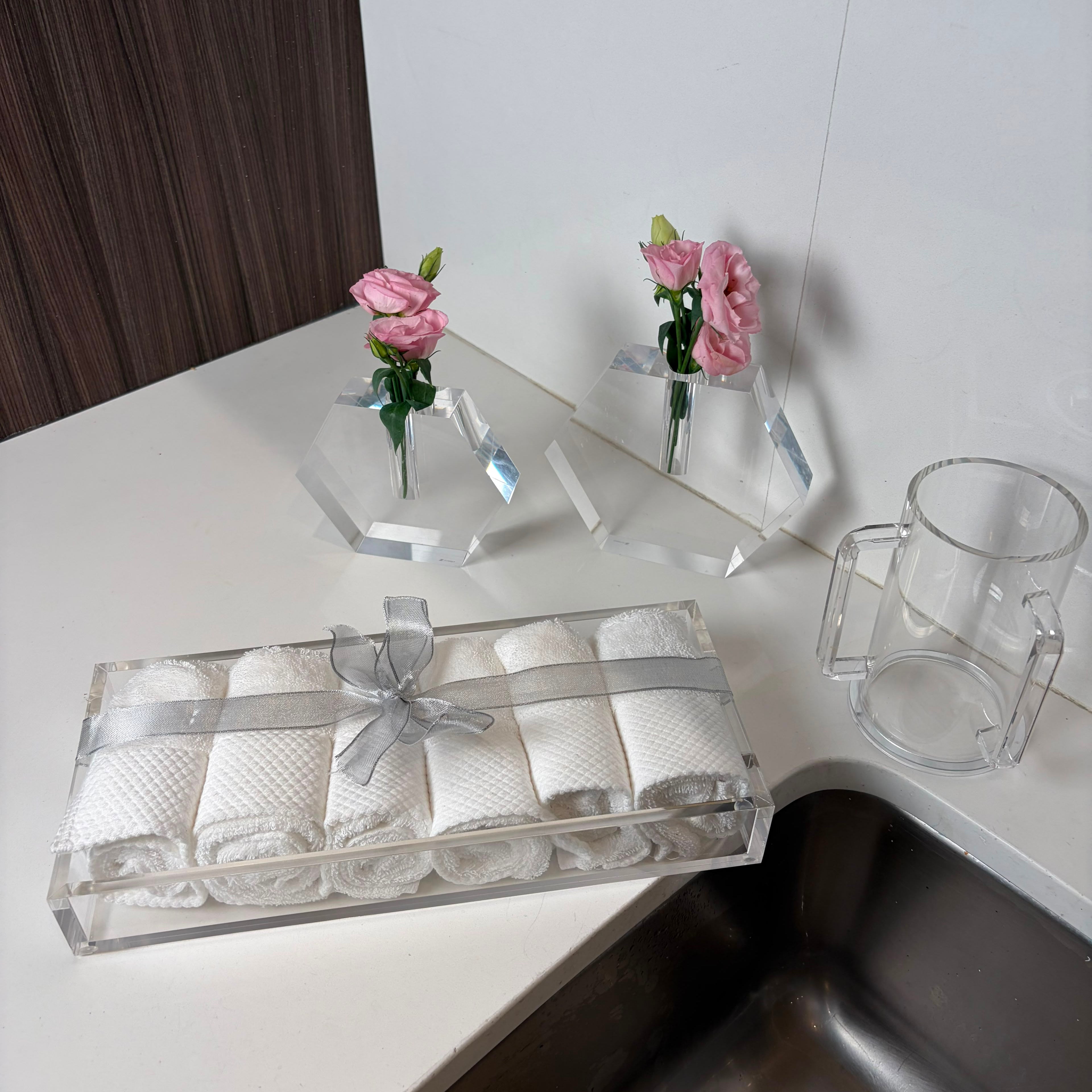 A clear acrylic gift box containing rolled white towels tied with a sheer gray ribbon, placed on a white countertop next to a stainless steel sink. Two small pink flowers in clear geometric holders and a clear lucite washing cup are also visible in the background.