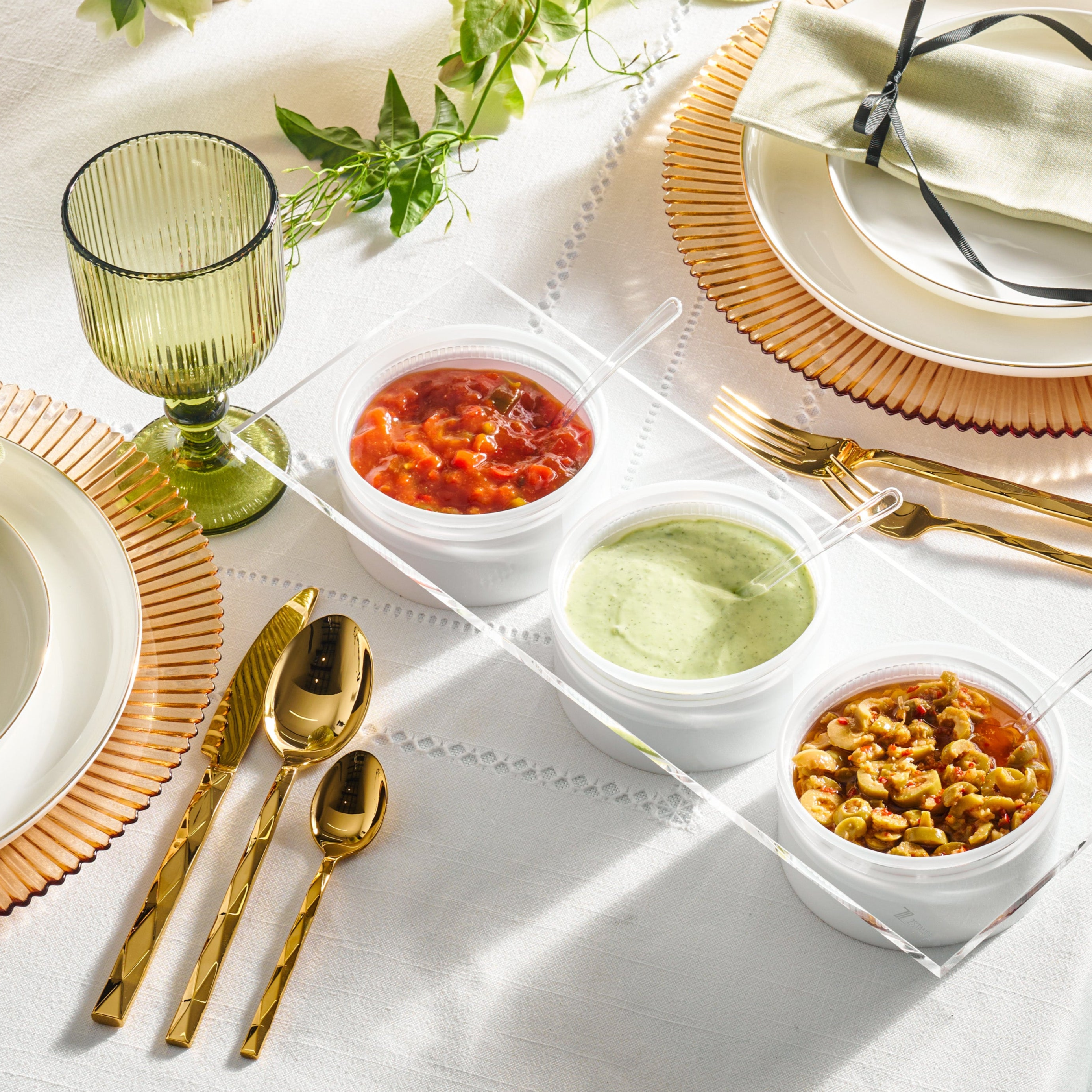 Table setting with gold cutlery, green glass, and small containers of food on a white tablecloth - Zstander