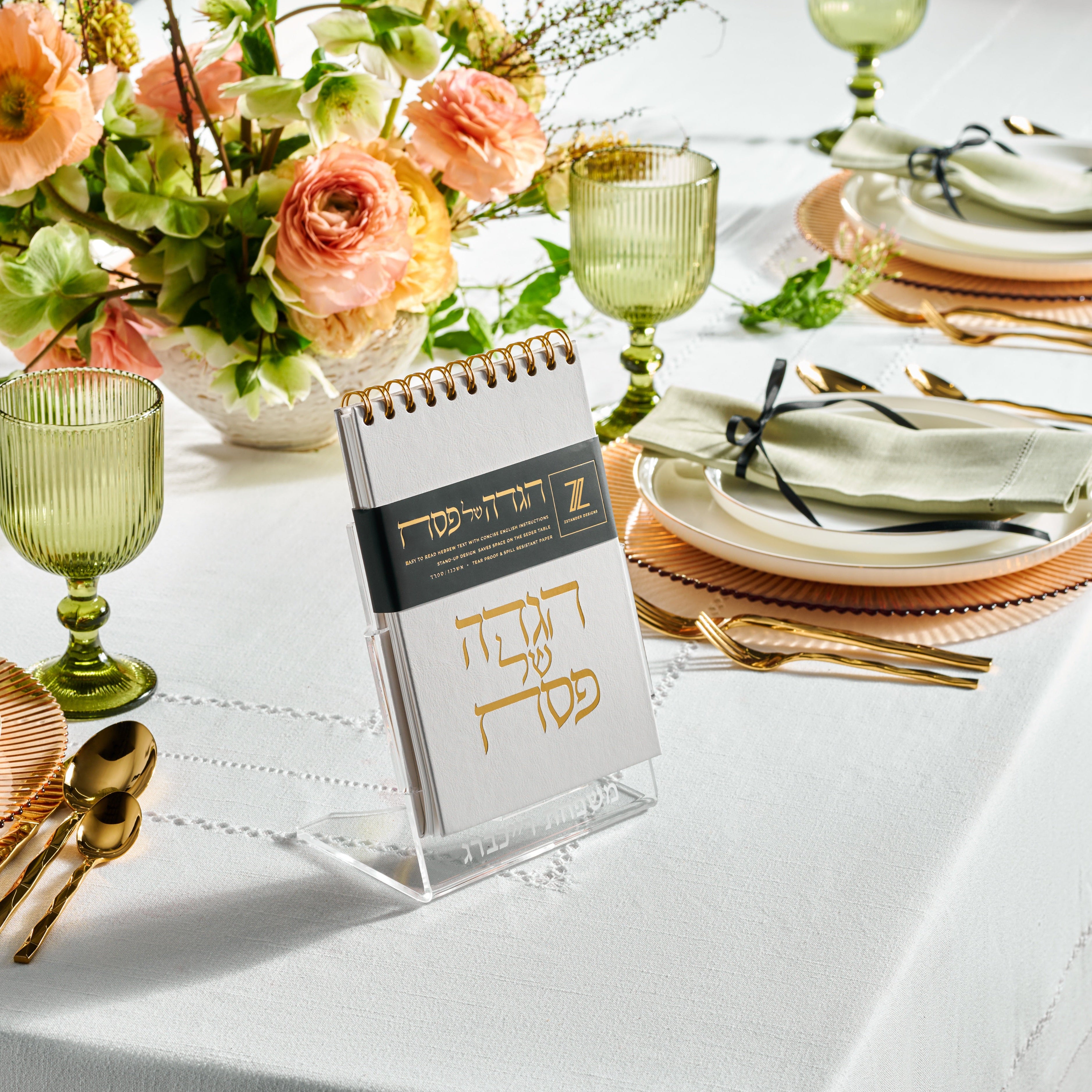 A spiral-top hagaddah on a lucite stand displayed on a set dining table with Seder plate, Kiddush cup, and decorative elements - Zstander