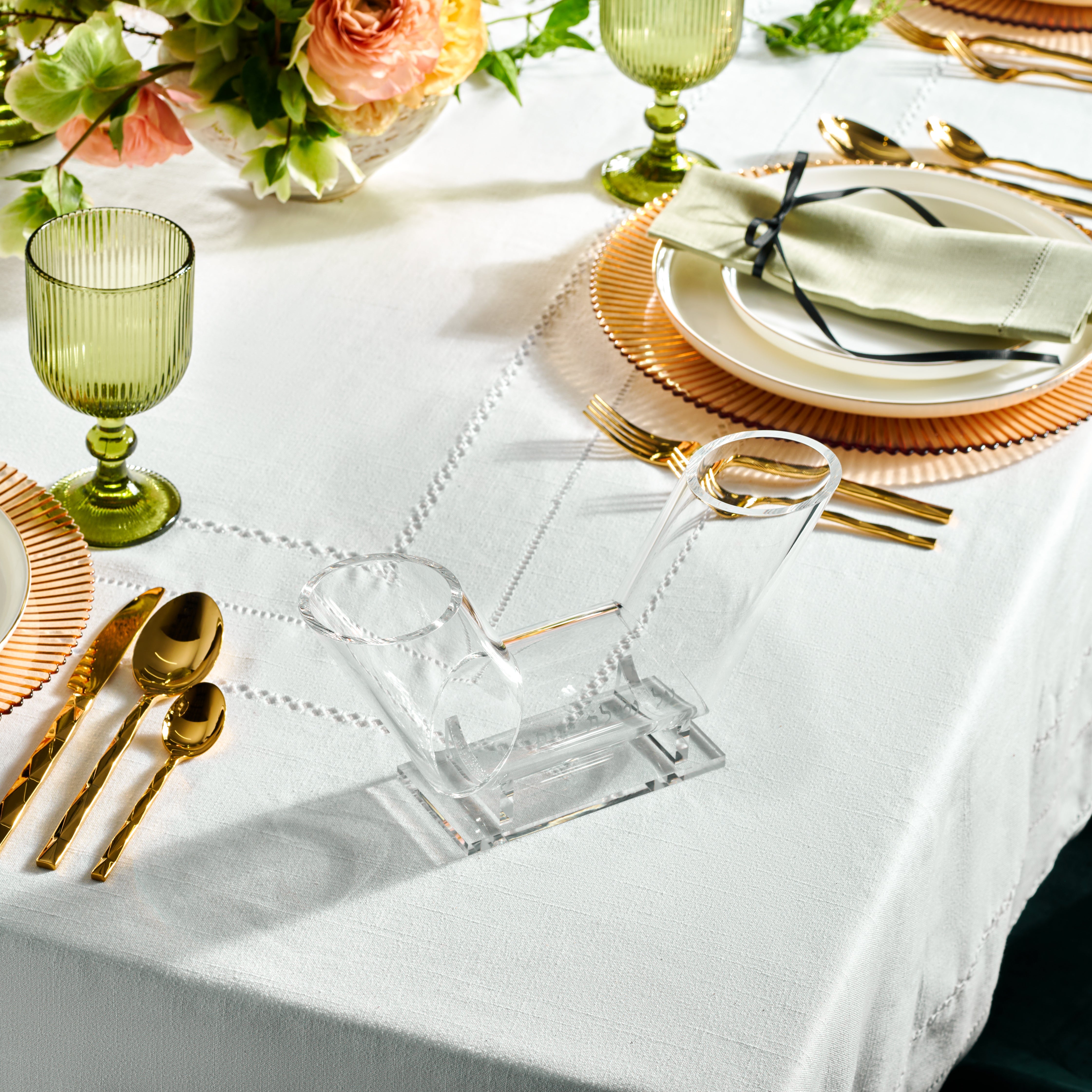 A clear Lucite dual spout washing cup placed on a white surface, likely a table, with elegant tableware in the background, including green glasses and gold flatware - Zstander