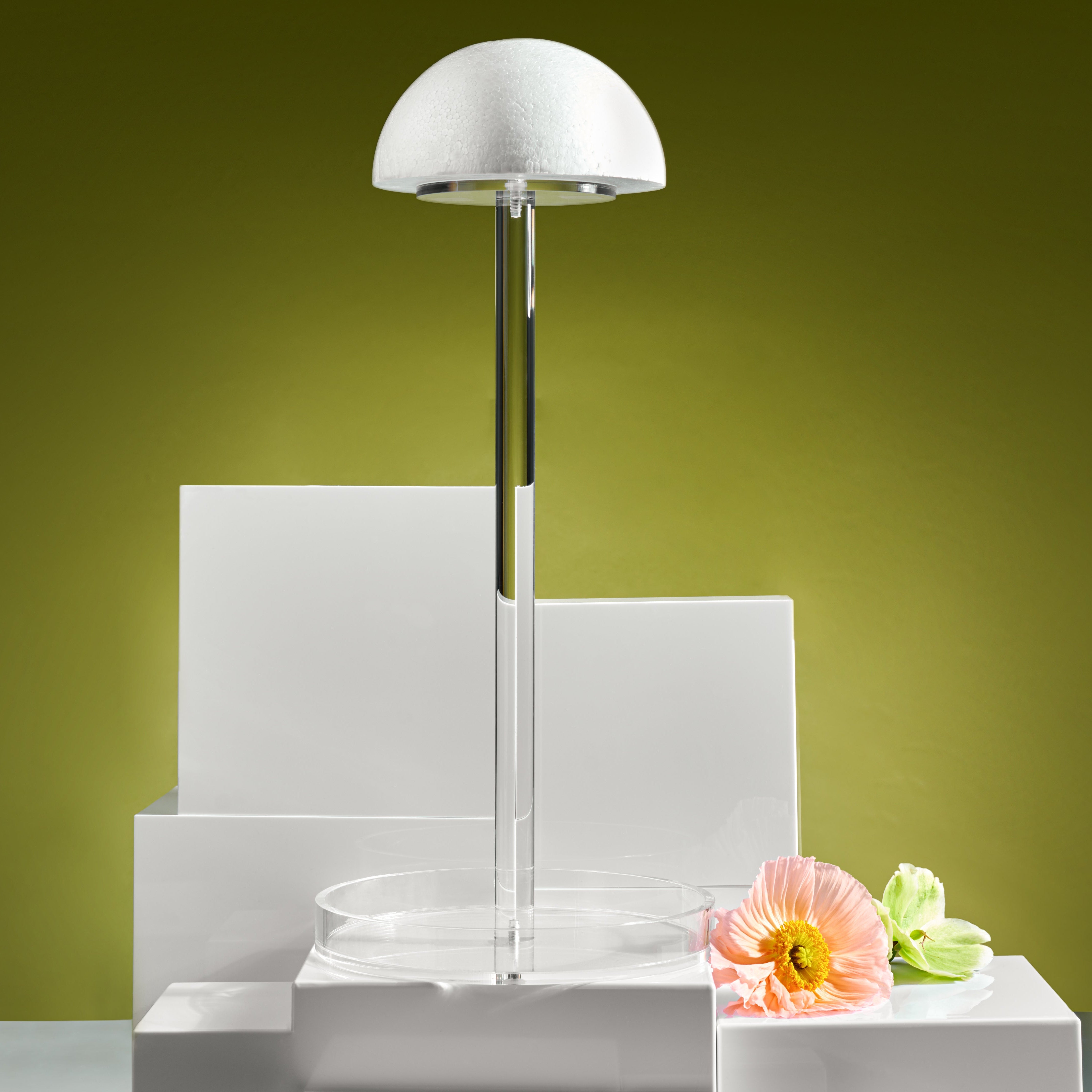 A clear acrylic wig stand with a round base, a vertical supporting rod, and a dome-shaped top, displayed against a yellow background with a few flowers at the base - Zstander