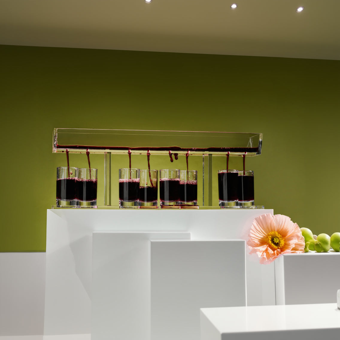 A clear acrylic wine glass fountain with eight glasses, arranged in a vertical line, with a green background and a pink flower in the foreground - Zstander