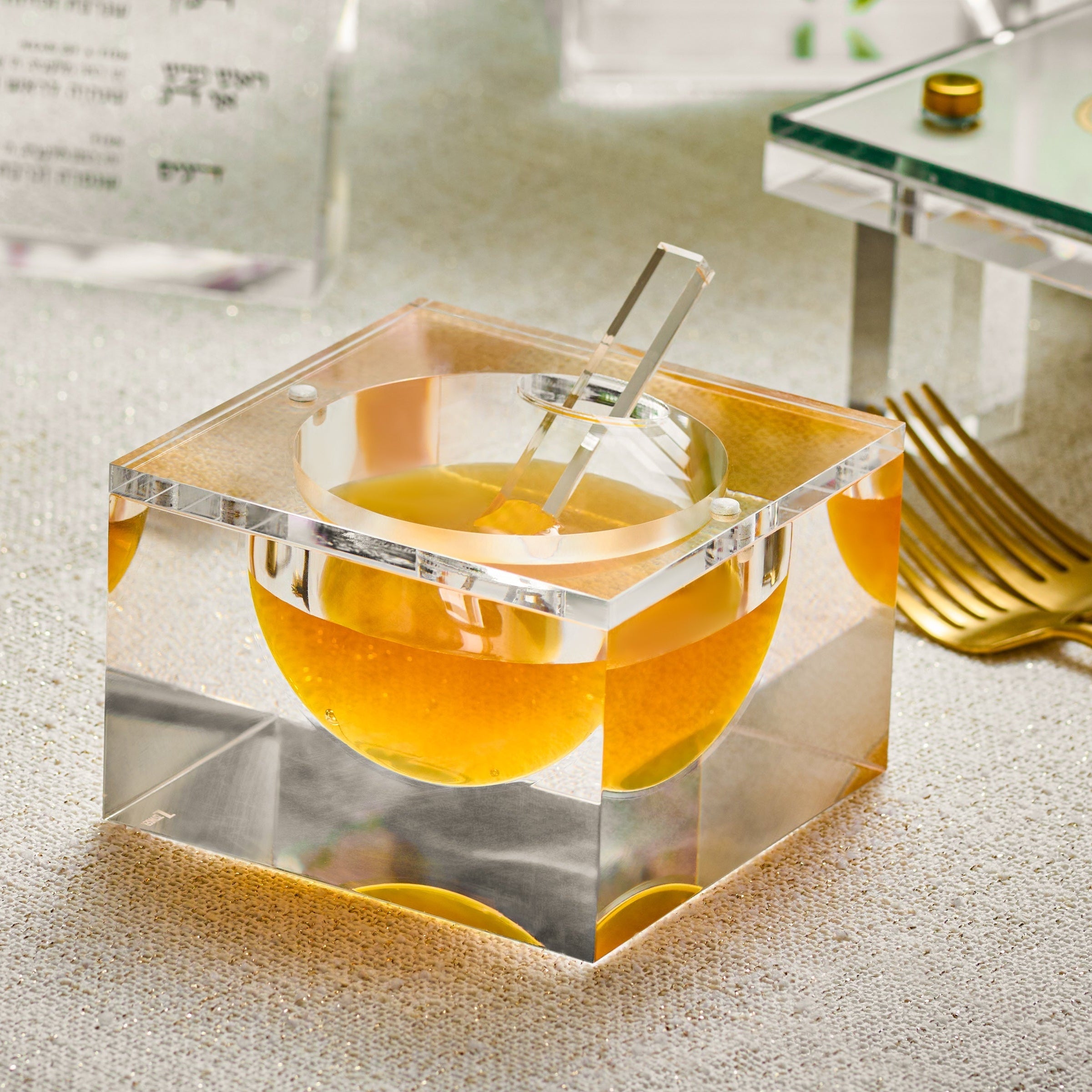 A clear acrylic square honey Dish holding honey, styled on a tabletop with gold-toned cutlery, flowers, and a glass menu card in the background - Zstander