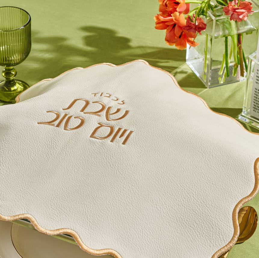 Table setting with a decorative challah cover, wine glasses, and flowers on a green tablecloth.