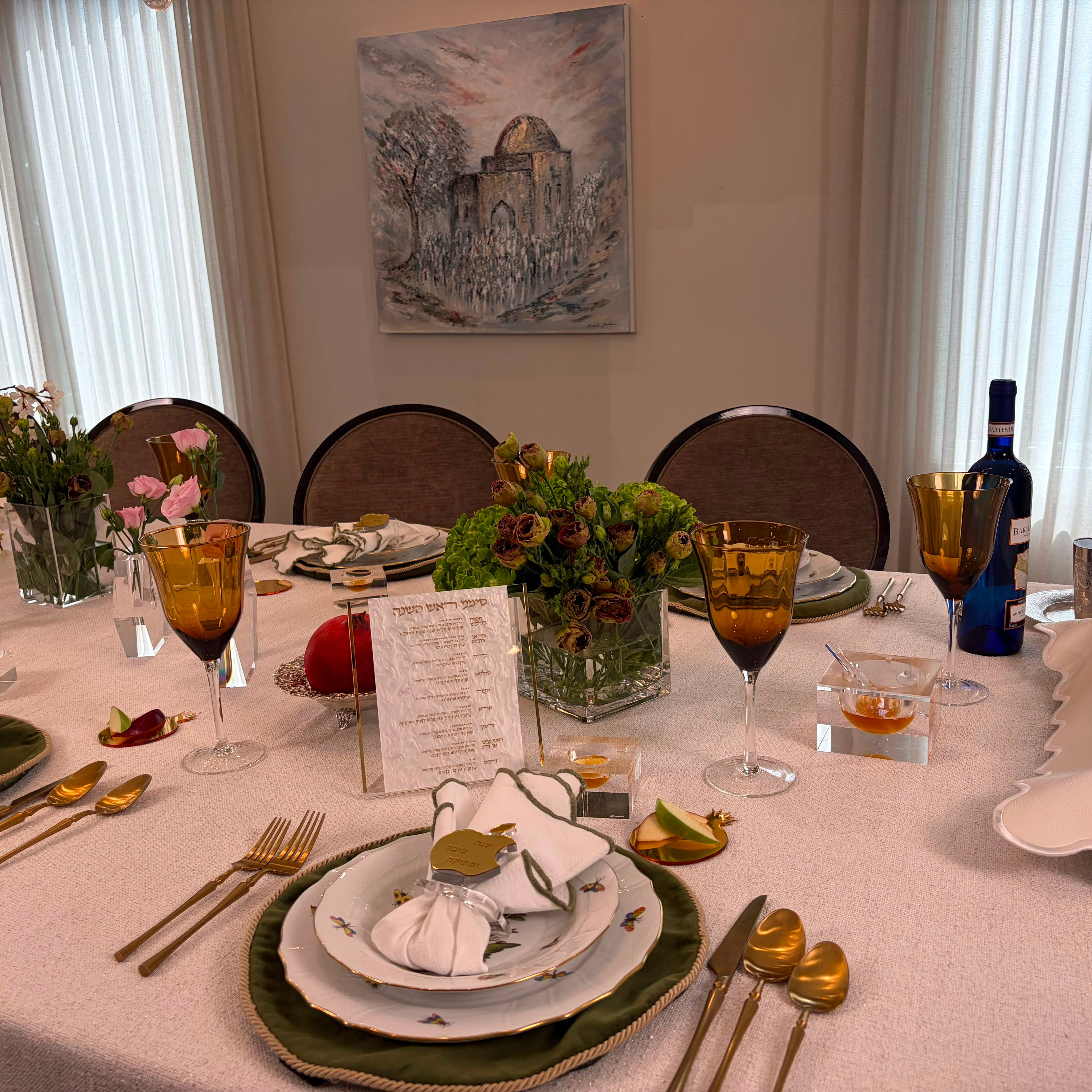 A beautifully set dining table in a dining room, featuring gold flatware, amber-tinted wine glasses, layered plates, floral centerpieces, and a white tablecloth. In the background, three dark wood chairs are positioned in front of a wall with a painting and white window curtains - Zstander