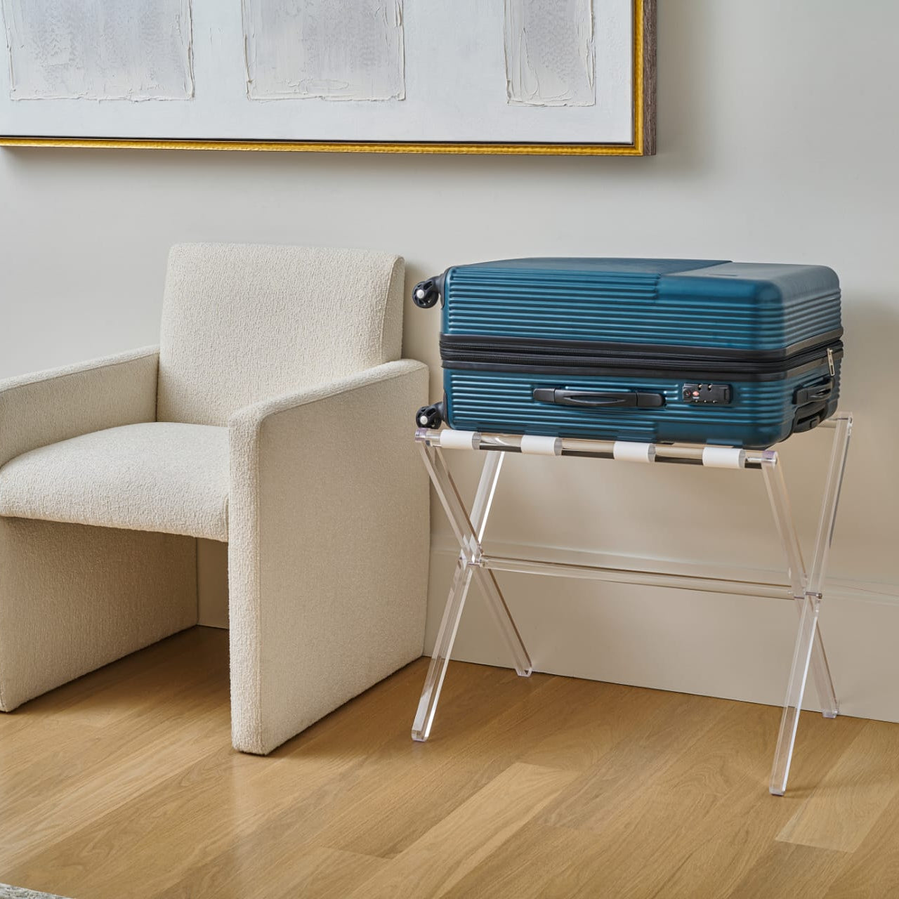 Blue suitcase on a luggage stand next to a beige armchair in a room with a framed picture on the wall - Zstander