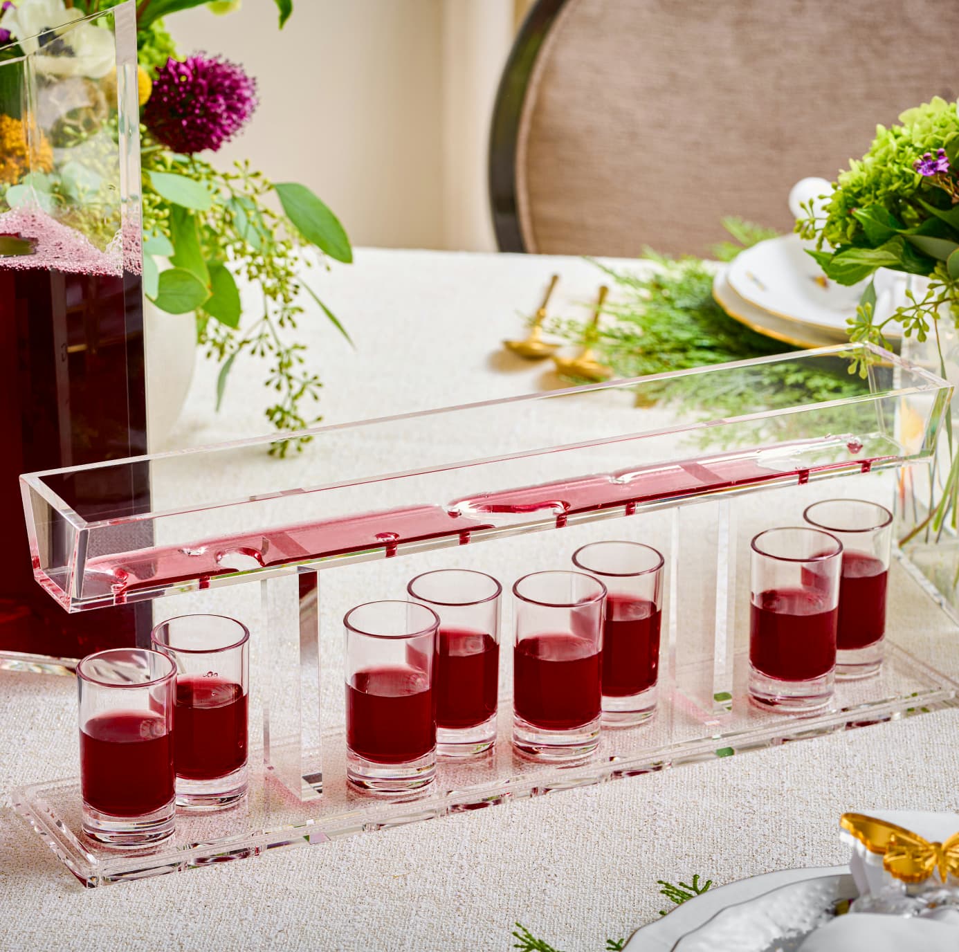 Row of shot glasses with red liquid on a clear tray, set against a blurred background with flowers and decor - Zstander