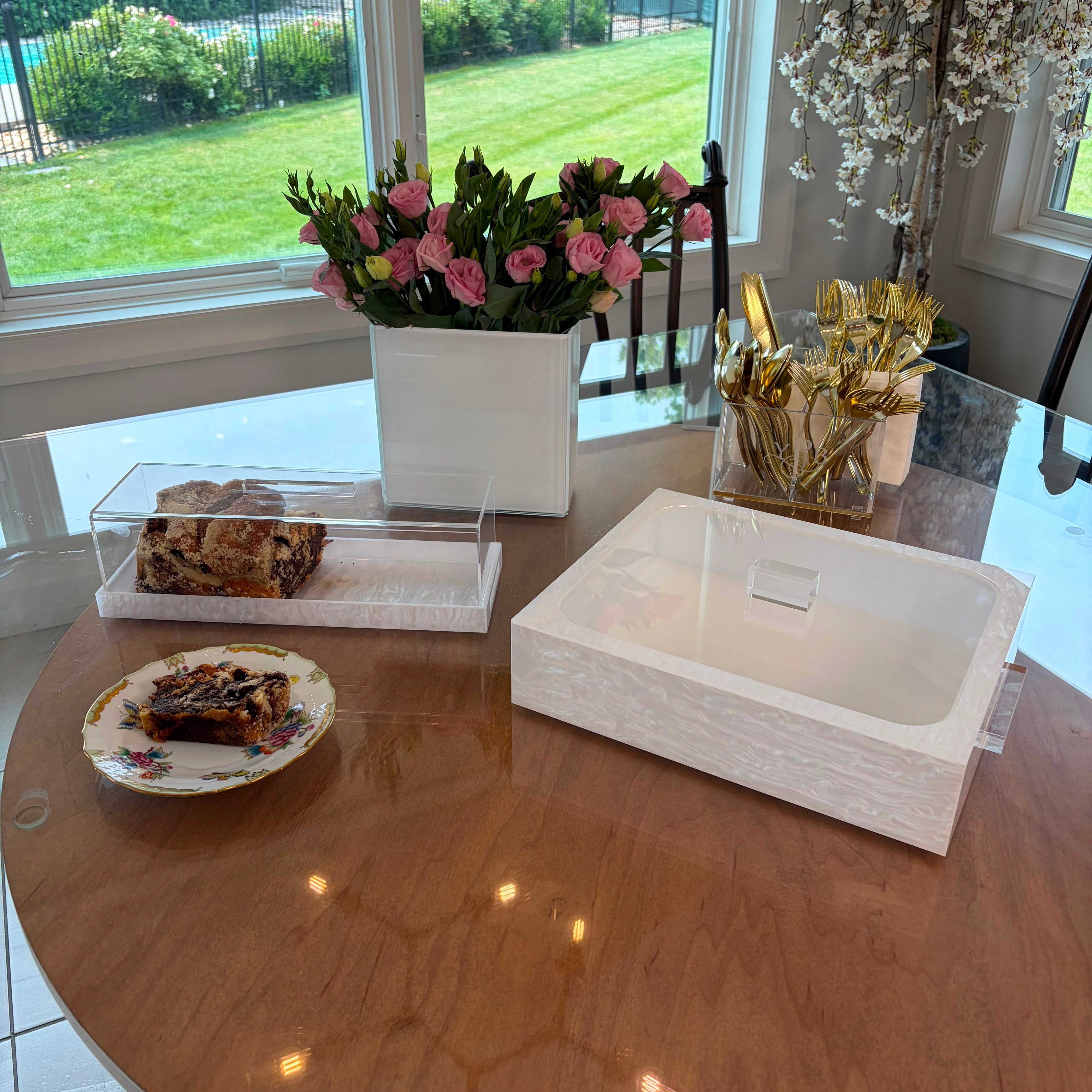 A white rectangular lucite cake tray sits on a round wooden table in a bright dining room. A clear plastic container with a tray holds a pastry, and another pastry is on a floral plate - Zstander