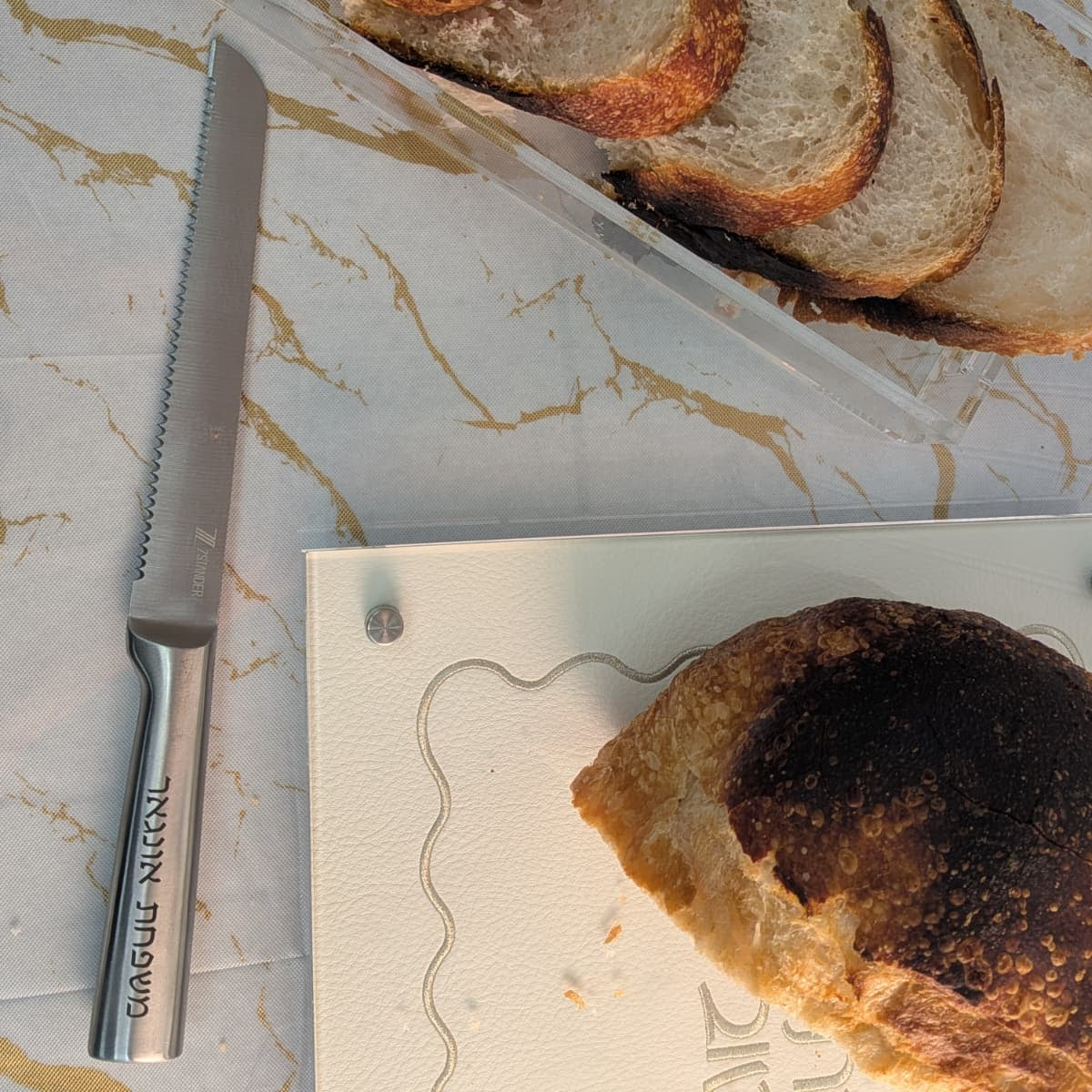 Sliced artisanal bread in a clear lucite tray on a marble-patterned surface, with a serrated bread knife and a white cutting board with a burnt piece of bread - Zstander