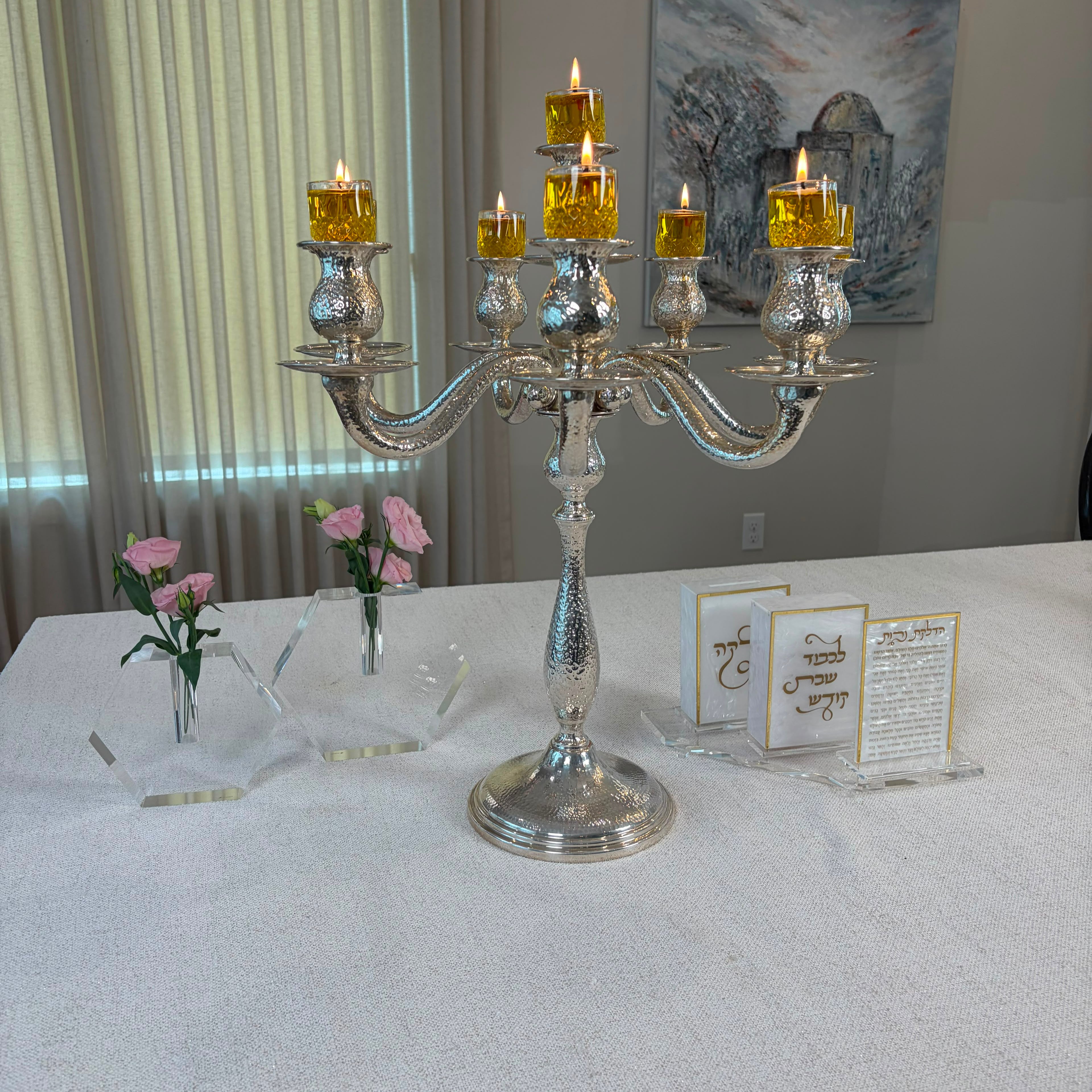 A silver candelabra with six lit yellow candles is displayed on a white tablecloth in an indoor setting, with small floral arrangements and printed cards nearby- Zstander