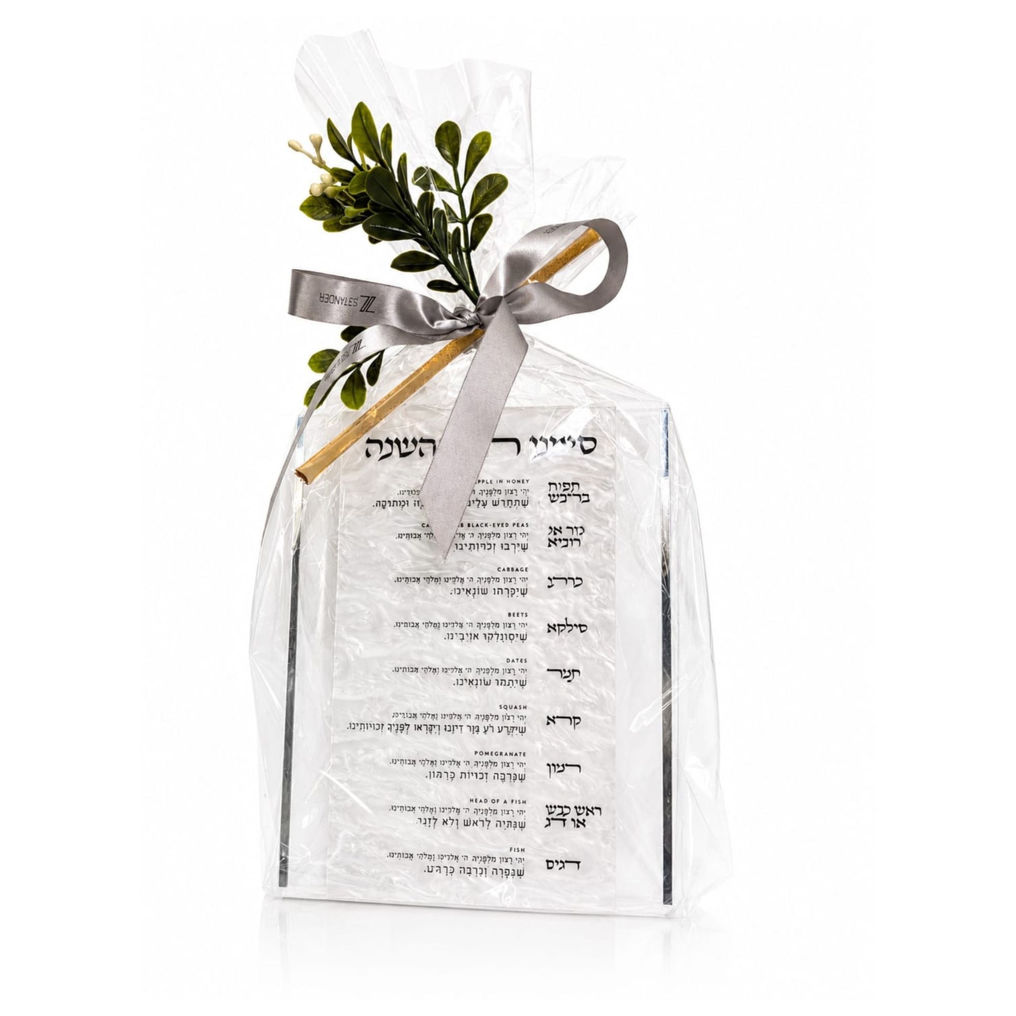 A clear cellophane-wrapped gift bag with a simanim plaque, tied with a silver ribbon and decorated with green leaves and a honey stick, shown on a white background - Zstander