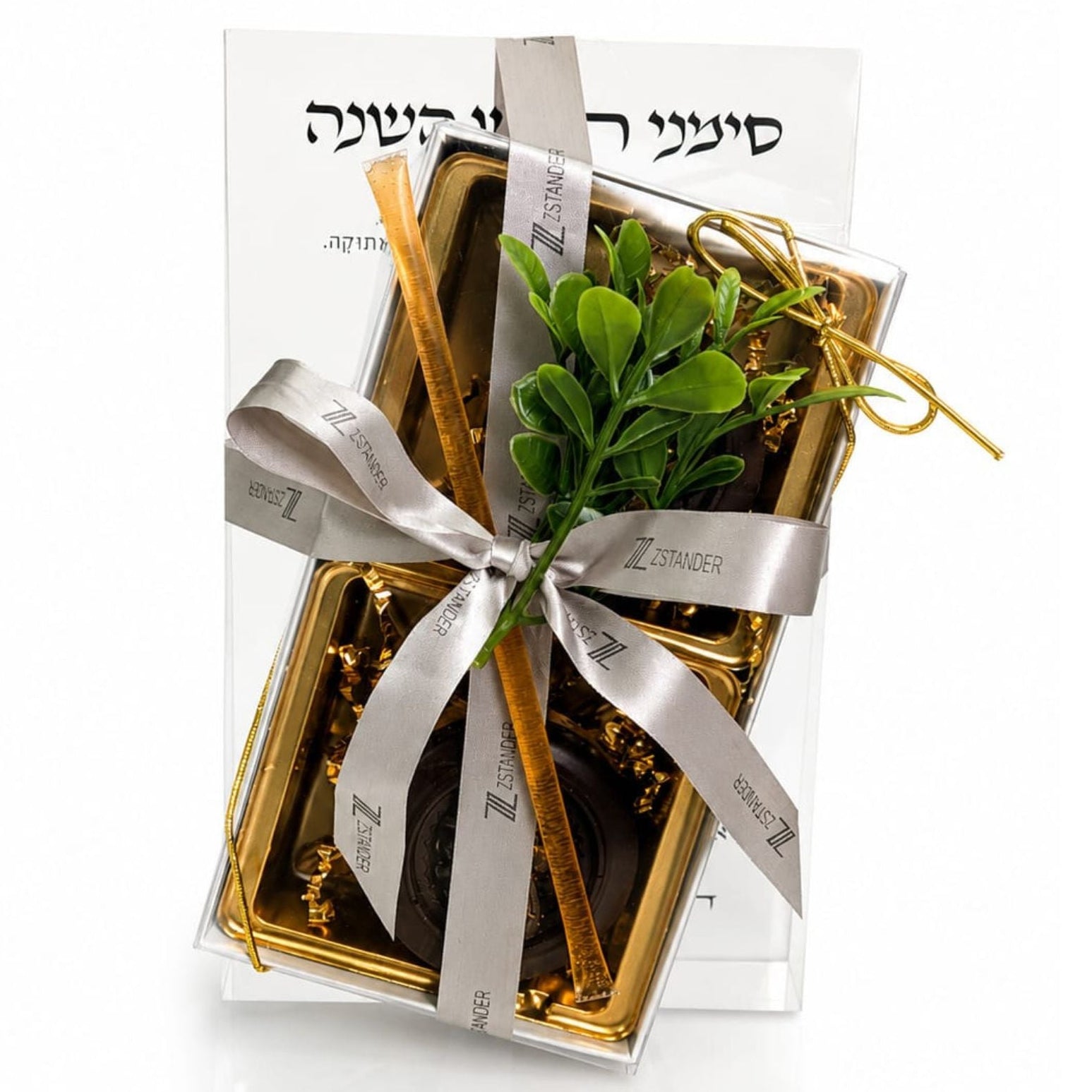 A gift box of chocolate and gold coins wrapped in a silver ribbon with a small green leaf sprig, presented on a white background. The box has Hebrew text on the top.
