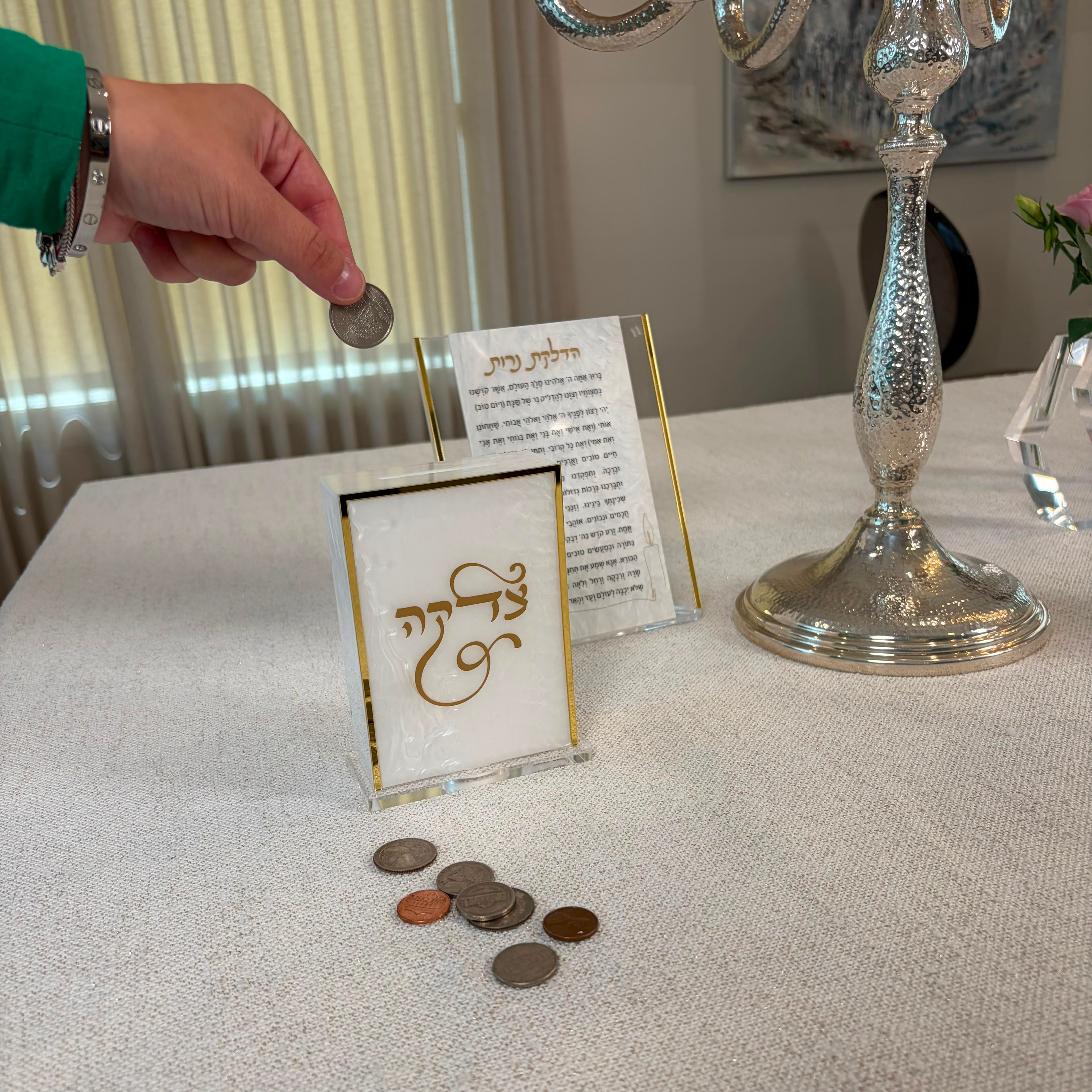 A hand drops a coin into a clear acrylic money box with gold trim on a beige tablecloth, with a silver candelabra and a small card in the background - Zstander