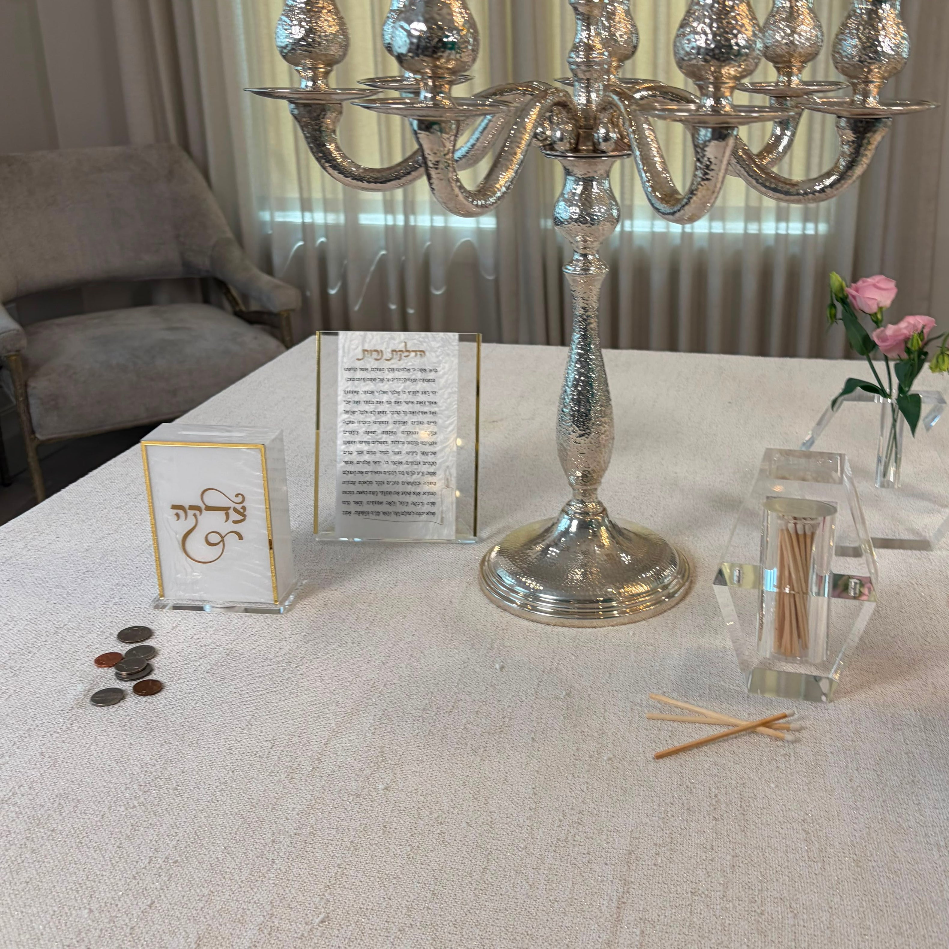 A silver candelabra with amber glass candle holders sits on a white tablecloth in a home setting, near a clear match box.