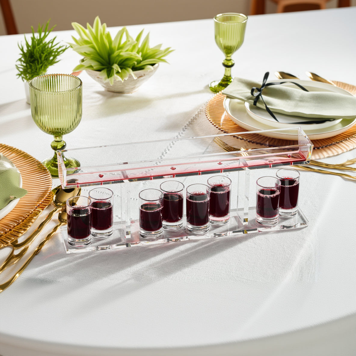 A clear lucite Kiddush fountain with multiple compartments, each filled with red liquid, likely wine, displayed on a table with tableware - Zstander