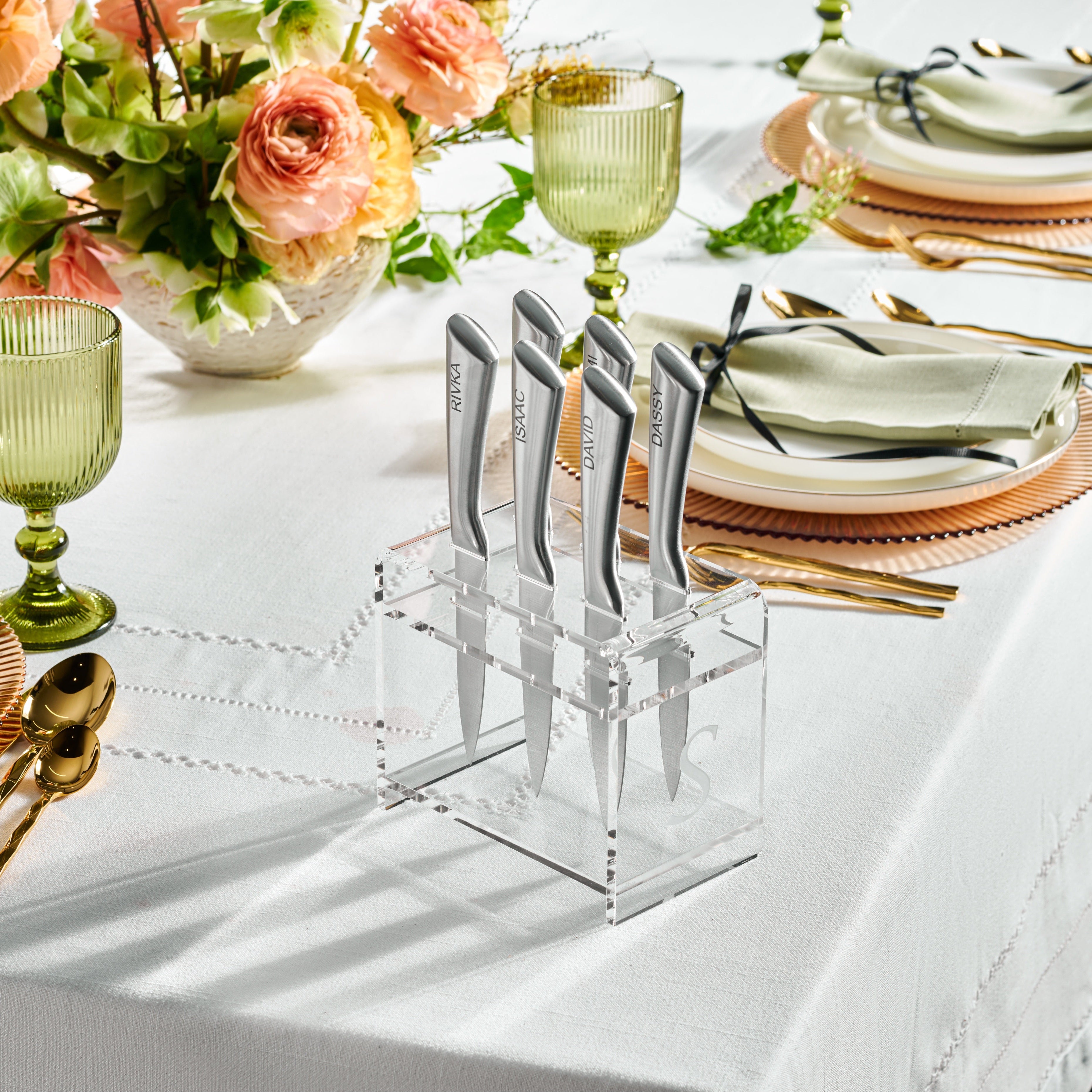 A set of silver steak knives placed in a clear lucite holder, displayed on a table set for dinner with plates, glasses, and flowers in the background - Zstander
