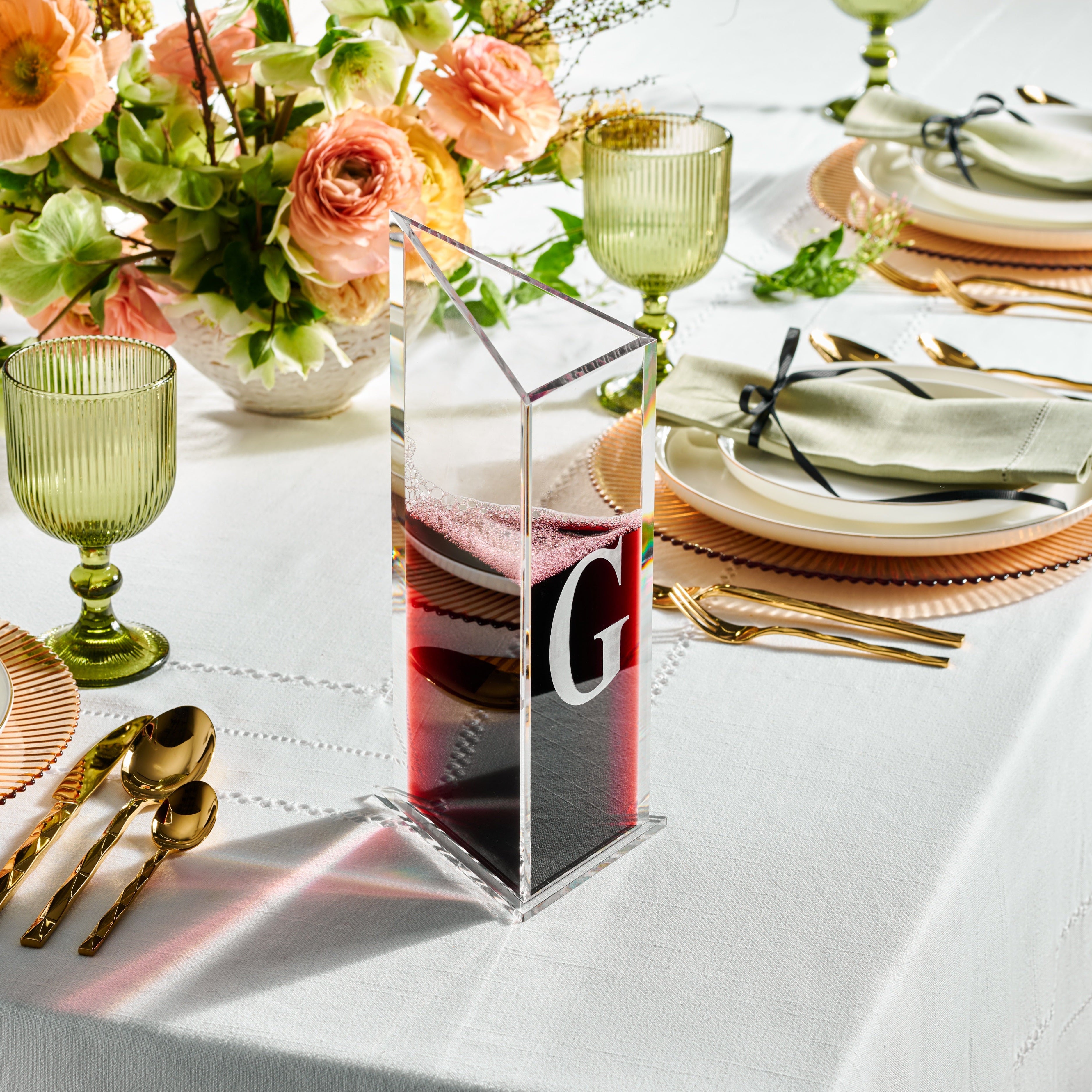 A clear lucite decanter with a geometric triangular shape, featuring a initial 'G' engraved on it, set on a table with a floral arrangement, green glasses, and diningware - Zstander