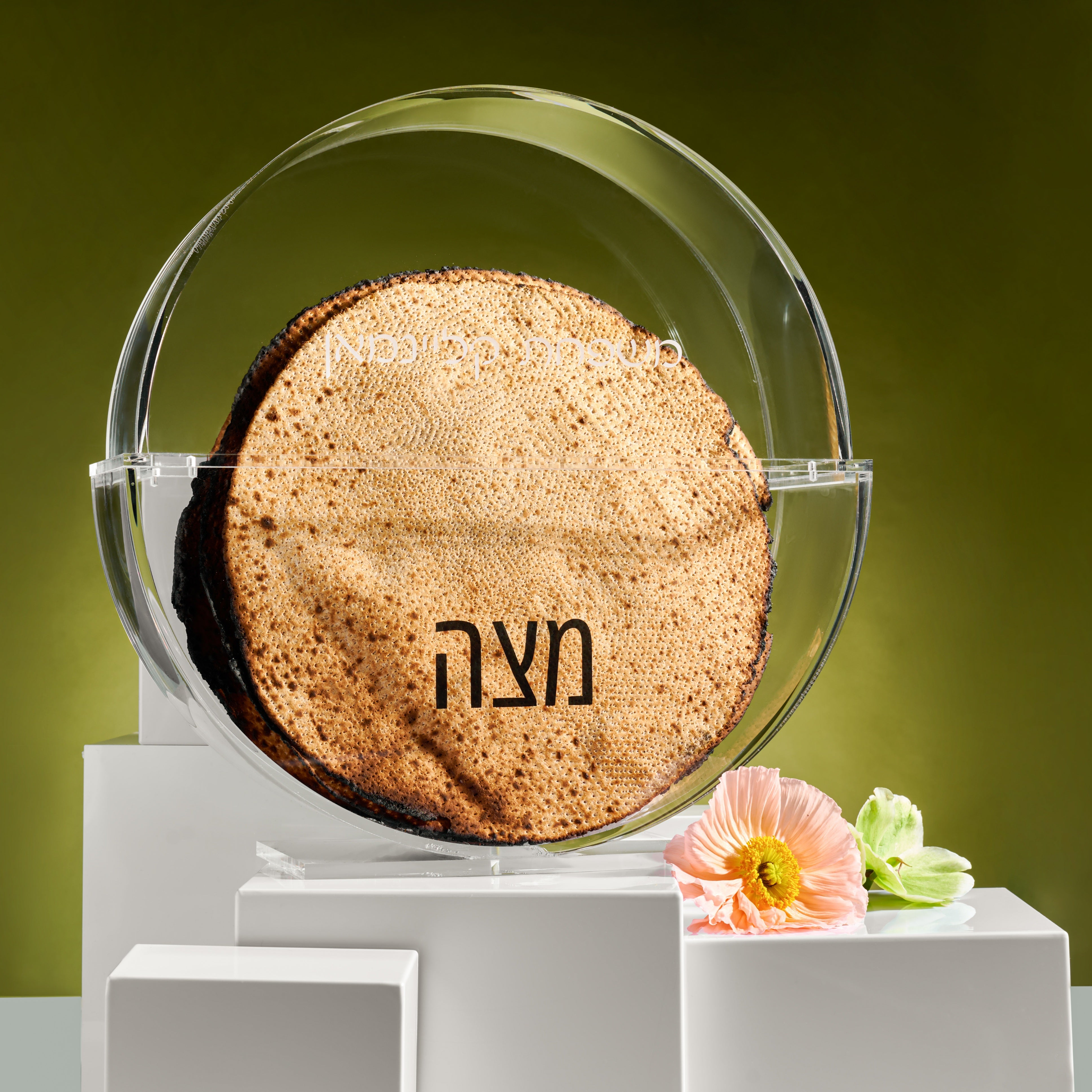 Round lucite matzah box with the words Matzah in hebrew on a clear stand against a green background - Zstander