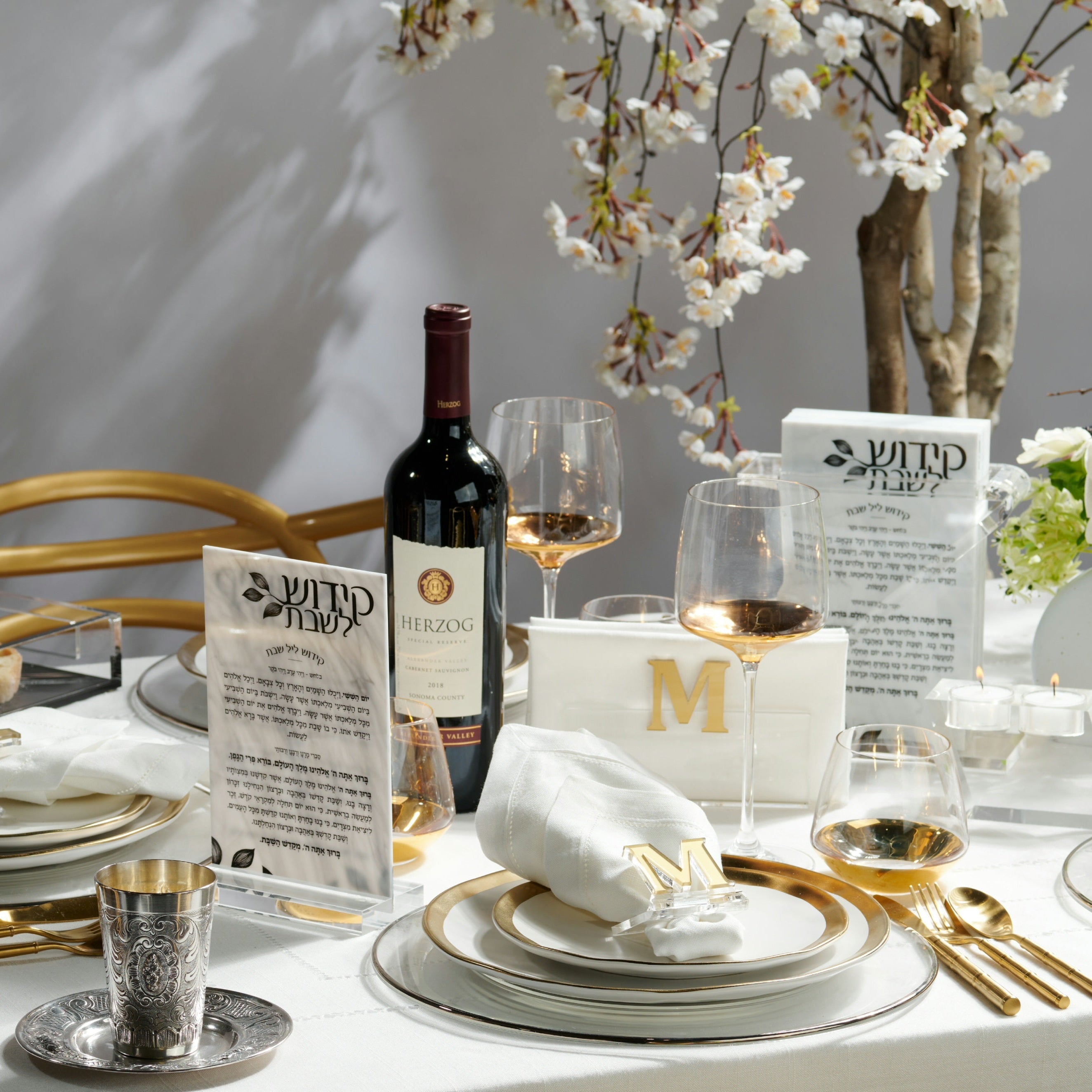 A beautifully set shabbos table featuring white plates with gold trim, white napkins with gold monogram letter accents, gold flatware, a bottle of Herzon red wine, clear wine glasses, and kiddush for shabbos under white cherry blossom branches- Zstander