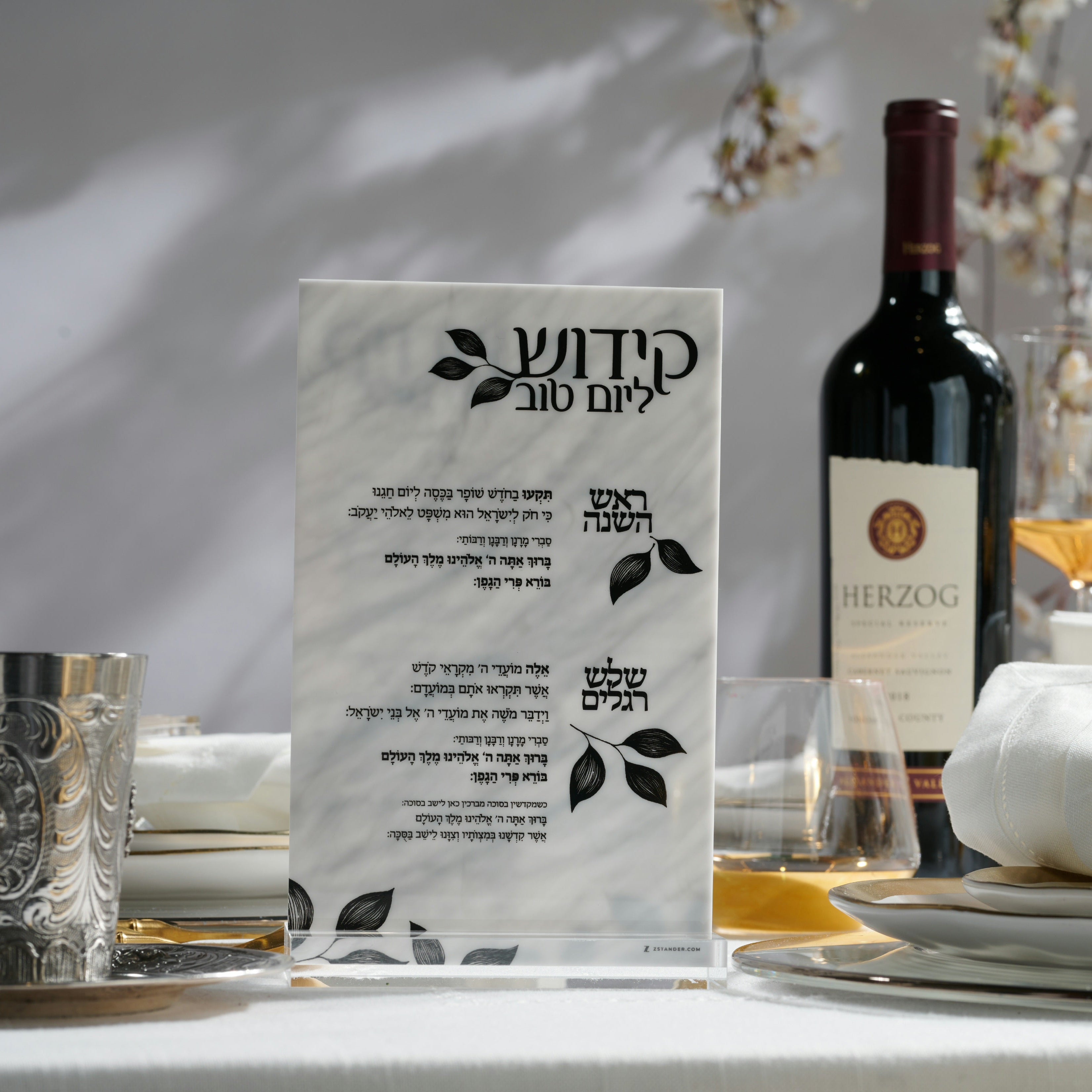A Kiddush card for shabbos in Hebrew stands on a table in a styled dining setting, with a bottle of red wine, glasses, plates, and flowers in the background - Zstander