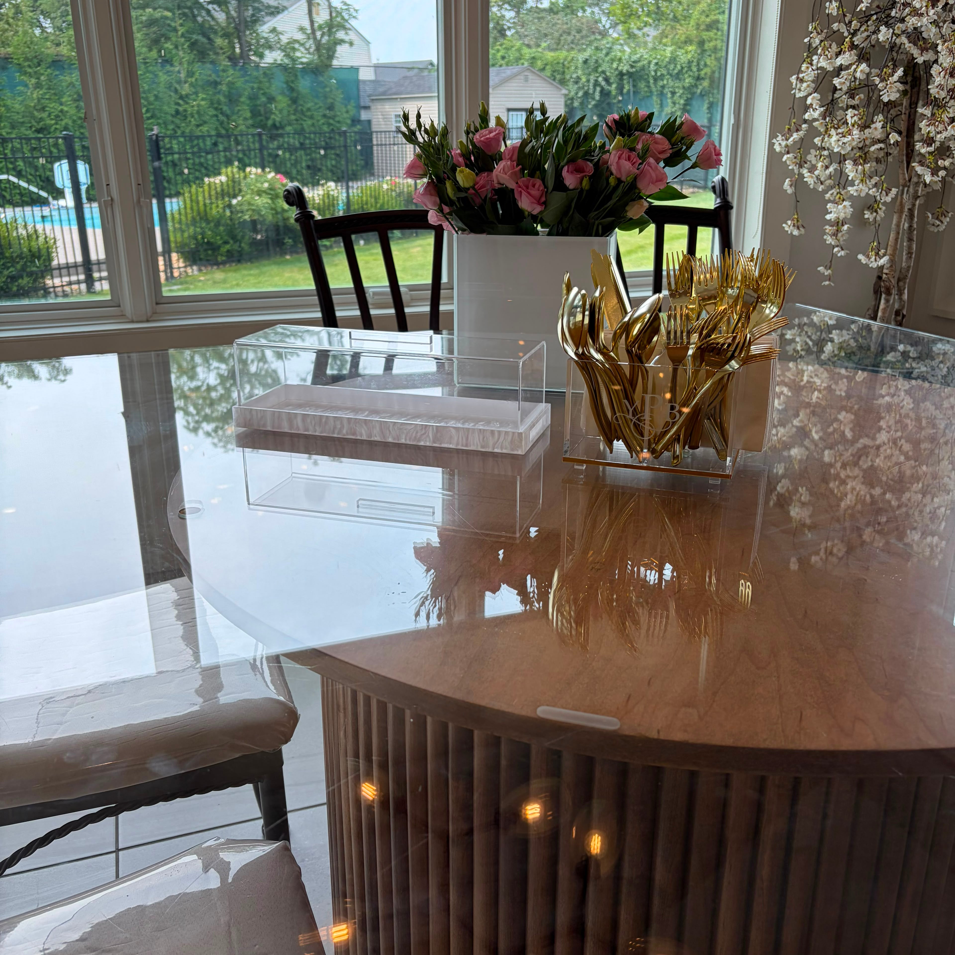 A modern curved dining table with a glossy light wood top and fluted slatted base, styled with a white vase of pink flowers and a gold utensil holder. The table is set in a bright room with large windows showing a backyard pool and trees.