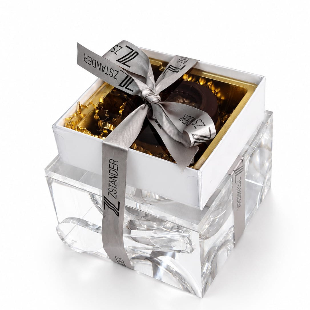 A square gift box with a clear lucite honey dish, topped with a white lid and wrapped in a silver ribbon printed with the brand name 'ZSTANDER'. The box is filled with gold crinkle paper and dark chocolates visible at the top - Zstander